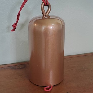 Może przedstawiać: Dzwonek w kolorze miedzi z czerwonym sznurkiem wiszącym z g&oacute;ry. Dzwonek ma cylindryczny kształt i gładkie, błyszczące wykończenie.