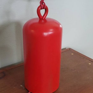 Może przedstawiać: Czerwony, cylindryczny dzwonek z pętlą do zawieszenia wykonaną z czerwonej sznur&oacute;wki. Dzwonek stoi na drewnianej powierzchni.