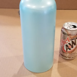 Może przedstawiać: Dzwonek ceramiczny w jasnoniebieskim kolorze z czarnym metalowym uchwytem i brązowym sk&oacute;rzanym paskiem. Dzwonek jest mniej więcej wielkości standardowej puszki napoju.