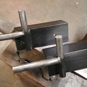 May include: Two black metal brackets with welded steel arms. The brackets are sitting on a metal anvil.