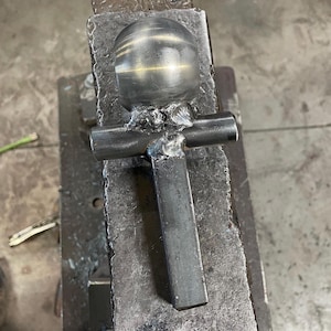 May include: A metal sculpture of a cross with a round ball on top. The sculpture is made of metal and has a rough, unfinished look. It is sitting on a metal surface.