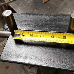 May include: A yellow measuring tape with black markings is placed across a black metal plate. The tape reads "1", "2", "3", "4", "5", "6", "7", and "8". There are two metal rods on the plate, one on each end.