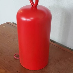 Może przedstawiać: Czerwony metalowy dzwonek z pętlą z sznurka wiszącą na g&oacute;rze. Dzwonek stoi na drewnianej powierzchni.