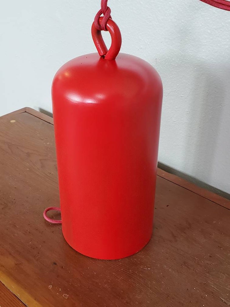 Może przedstawiać: Czerwony metalowy dzwonek z pętlą z sznurka wiszącą na g&oacute;rze. Dzwonek stoi na drewnianej powierzchni.
