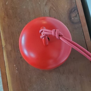 Może przedstawiać: Czerwony ceramiczny dzwonek z czerwonym sznurkowym uchwytem. Dzwonek leży na drewnianej powierzchni.