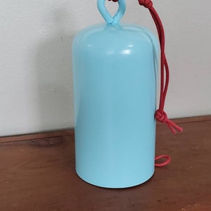 Może przedstawiać: Dzwonek metalowy w jasnoniebieskim kolorze z czerwonym sznurkowym uchwytem. Dzwonek jest zawieszony na drewnianej powierzchni.