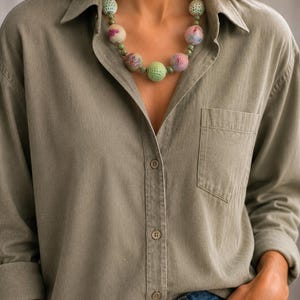 May include: A necklace featuring a series of round beads, including several light green crocheted spheres and larger felted balls in shades of pink, white, and blue, interspersed with small, smooth, dark green accent beads on a cord.