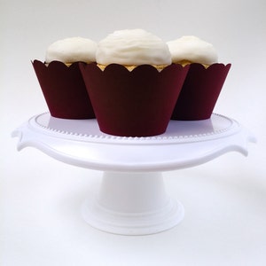 May include: Three white cupcakes with burgundy cupcake liners on a white cake stand.