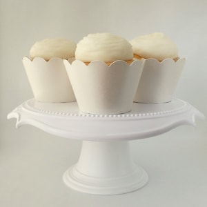 May include: Three white cupcakes with white frosting on a white cake stand. The cupcakes are in white paper liners with scalloped edges.