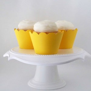 May include: Three yellow cupcake liners with white frosting on top of cupcakes. The cupcakes are on a white cake stand.