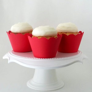 May include: Three white frosted cupcakes with red cupcake liners on a white cake stand.