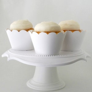 May include: Three white cupcakes with white frosting are displayed on a white cake stand. The cupcakes are in white scalloped cupcake liners.