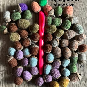 May include: A collection of colorful felt acorns in various shades of brown, green, blue, purple, and orange. A pink highlighter is placed vertically to show size. The acorns have brown caps and are arranged on a wooden surface.