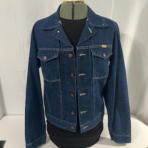May include: A dark blue denim jacket with a button-up front, two chest pockets, and two side pockets. The jacket has a classic collar and is displayed on a mannequin. The jacket has a small, rectangular tag on the right chest.