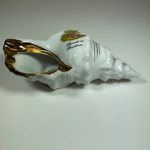 May include: White porcelain seashell with gold trim and a painted floral design. The shell has the words "Recuerdo de Benidorm" written on it.
