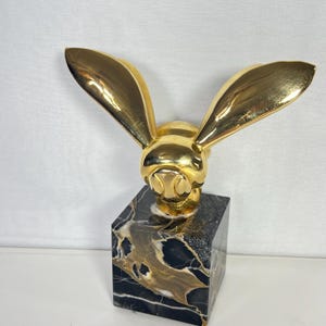 May include: A gold-colored rabbit sculpture with long, upright ears. The rabbit head is mounted on a black and gold marble cube base. The sculpture is a decorative item.