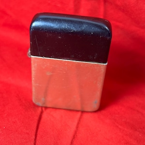 May include: A vintage lighter with a rectangular shape. The top is black, and the bottom is a metallic orange color. The lighter has a hinged lid and a silver metal accent. The lighter is set against a red background.