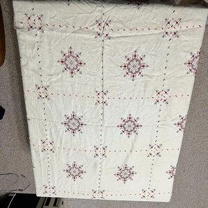 May include: Cream-colored textile with a repeating pattern of red and black cross-stitch floral designs. The fabric appears to be linen or a similar natural fiber, with a slightly textured surface. The overall design is symmetrical and geometric.