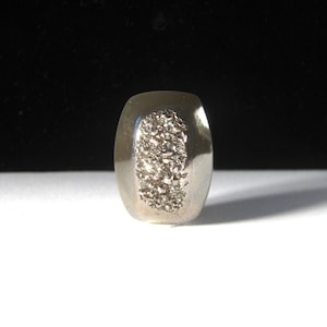 Puede incluir: Un anillo plateado con forma rectangular y bordes redondeados. El centro del anillo presenta un diseño cristalino texturizado. El anillo está sobre una superficie blanca con un fondo negro.