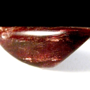 May include: A polished, dark red gemstone with a curved, boat-like shape. The gem has a smooth, reflective surface with internal inclusions, set against a white and black background.