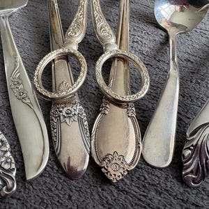 May include: Close-up of antique silverware with floral and ornate designs. Includes a cake server, serving spoons, and sugar tongs. The silver utensils are arranged on a grey surface.