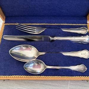 May include: A vintage silver cutlery set, comprising a fork, knife, and two spoons, displayed in a brown leatherette case with a blue velvet lining. The silverware has decorative handles.