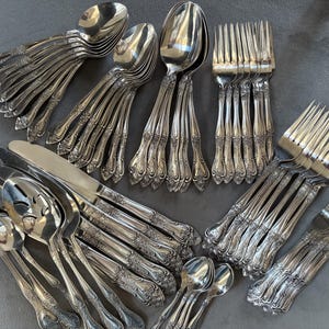 May include: An assortment of silver-colored flatware, including spoons, forks, knives, and serving spoons, arranged on a gray surface. The silverware has ornate, detailed handle designs. The arrangement suggests a complete set for dining.