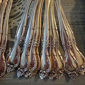 May include: Close-up of a collection of antique silver forks. The forks have ornate floral designs on the handles and tines. The silver forks are arranged in a fan shape, reflecting light. The forks are on a dark gray surface.