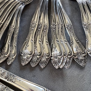 May include: Close-up of antique silverware with floral detailing. The silver-coloured forks, spoons, and knives are arranged on a grey surface. The intricate floral pattern adds a touch of elegance to the vintage flatware.