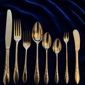 May include: A set of eight silver-toned metal utensils arranged in a row, including a dinner knife, two forks of varying sizes, three spoons of different sizes, a small cocktail fork, and a butter knife, all featuring a leaf motif on the handles.