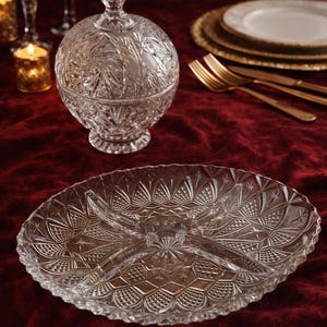 May include: A clear, cut-glass serving platter with a divided design and a matching lidded candy dish. The glassware features intricate starburst and geometric patterns. Gold-toned flatware and plates are visible in the background, set on a deep red velvet tablecloth.