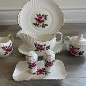 Puede incluir: Un juego de té de porcelana blanca con un diseño de rosa rosa. El juego incluye una crema, azucarero, salsera, plato y saleros y pimenteros. Cada pieza tiene un borde dorado y una textura acanalada.
