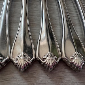 Peut inclure: Gros plan sur une collection de couverts argentés. Les couverts présentent un motif floral décoratif à la base du manche. Les manches sont orientés vers le spectateur et les couverts sont disposés en rangée.
