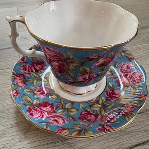 May include: A blue and white teacup and saucer set with a floral pattern of pink roses. The cup and saucer have a gold trim.