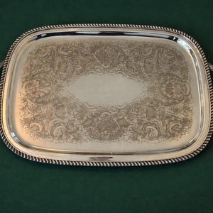 May include: A rectangular silver serving tray with ornate floral and scrollwork designs. The tray has a rope-like border and curved handles on each side. The tray is displayed on a dark green surface.