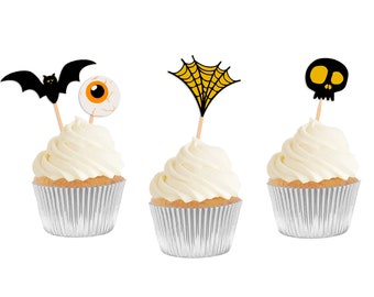 May include: Three cupcakes decorated with Halloween themed toppers. The cupcakes are frosted with white frosting and have silver cupcake liners. The toppers are a black bat, a yellow spiderweb, and a black skull with yellow eyes.