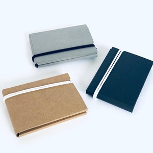 Puede incluir: Tres pequeños cuadernos con cierres elásticos. Los cuadernos están hechos de diferentes materiales: gris, marrón y negro.
