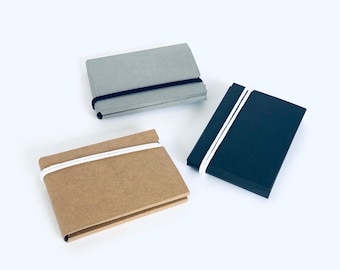 May include: Three small notebooks with elastic closures. The notebooks are made of different materials: gray, brown, and black.