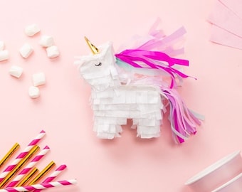 May include: A white unicorn pinata with pink and purple streamers on a pink background. The pinata has a gold horn and closed eyes. There are white marshmallows and pink and gold striped straws in the image.