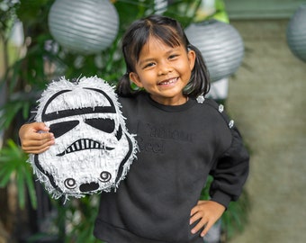 May include: A young girl smiles while holding a white and black Star Wars Stormtrooper piñata. She is wearing a black sweatshirt with the text "L'amour Reel" on the front.