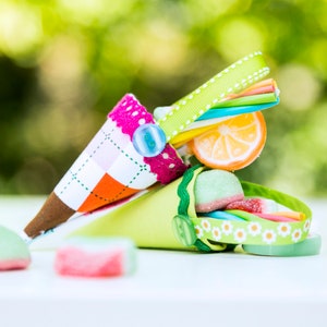 Puede incluir: Dos conos de papel coloridos llenos de caramelos, incluyendo caramelos gomosos, piruletas y un caramelo con sabor a cítricos. Los conos están decorados con cinta y botones.
