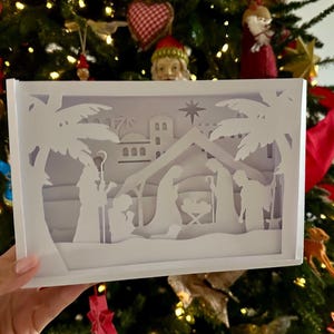 Puede incluir: Una obra de arte en papel blanco en capas que representa la escena de la Natividad. La obra presenta siluetas de María, José, el niño Jesús y otras figuras, sobre un fondo de un establo y palmeras. La pieza se sostiene frente a un árbol de Navidad.