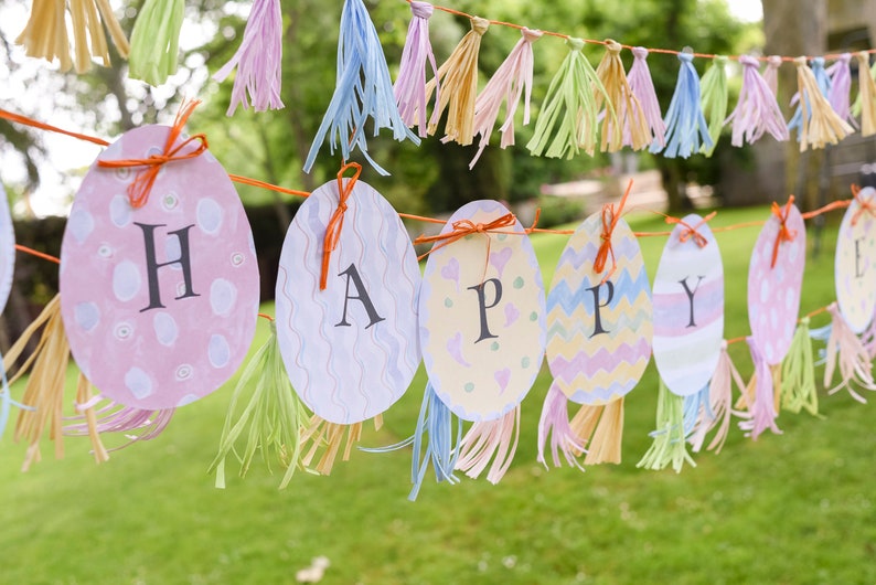 May include: A colorful banner with the word "HAPPY" spelled out on pastel-colored egg shapes. Each egg has a different pattern and is decorated with orange ribbon. The banner is strung with light blue, pink, and yellow tassels.