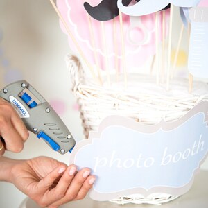 May include: A person is using a glue gun to attach a light blue and brown sign that says "photo booth" to a white wicker basket. The basket is filled with various paper photo booth props, including a mustache, a baby bottle, and a pink bow.
