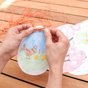 May include: A person is tying a string around a paper Easter egg with a picture of a hen and chicks. The egg is blue and white with a green background. There are other Easter eggs in the background.