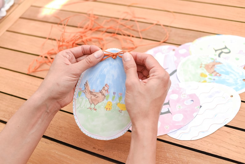 May include: A person is tying a string around a paper Easter egg with a picture of a hen and chicks. The egg is blue and white with a green background. There are other Easter eggs in the background.
