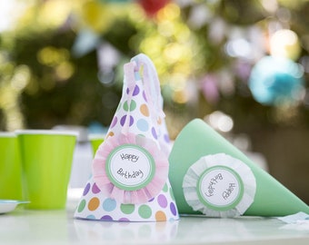May include: Two colorful paper party hats with white polka dots and a green and white "Happy Birthday" label. The hats are on a white table.