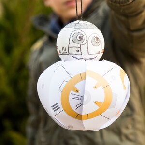 May include: A white and gold paper craft of a droid character from the Star Wars franchise. The droid has a round head with a face drawn on it and a round body with a gold circle design.