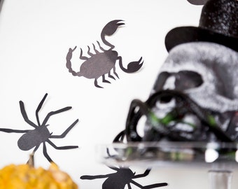 May include: Black paper cutouts of a spider, scorpion, and a skull with a top hat. The skull is out of focus and the cutouts are on a white background.