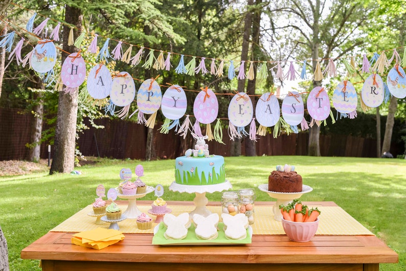 May include: An Easter party table setting with a green and blue cake decorated with a bunny, cupcakes, cookies, and a chocolate cake. The table is decorated with a yellow runner and a banner that says "Happy Easter".